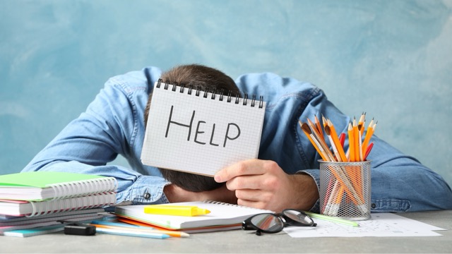 Un élève posant la tête sur un bureau avec un cahier affichant "aide" entouré de fournitures scolaires, révélateur d'un stress d'orientation scolaire.