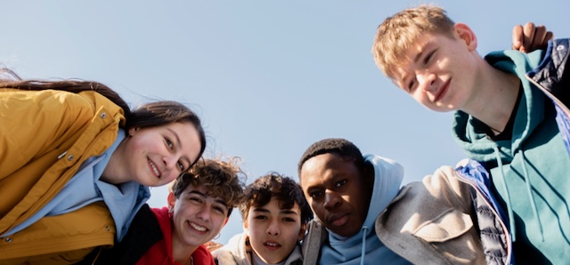 Cinq jeunes amis souriants et penchés ensemble pour une photo de groupe sous un ciel bleu clair.