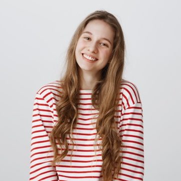 Jeune femme aux cheveux longs portant un pull rayé rouge et blanc, souriant à la caméra sur un fond blanc.